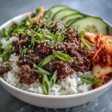 Spicy Korean Beef Bowl topped with crisp cucumber, tangy kimchi, and sesame seeds, served on a white plate.