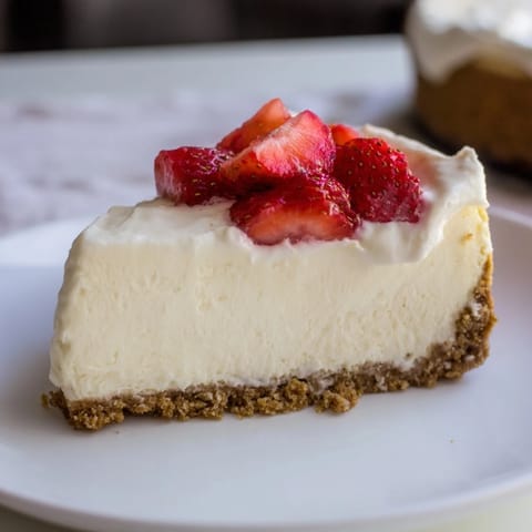 A thick slice of creamy New York Cheesecake with a graham cracker crust sits on a white plate.