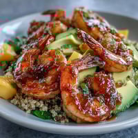 Grilled shrimp and creamy avocado slices rest atop fluffy quinoa, garnished with vibrant mango salsa and a drizzle of zesty lime chili sauce.