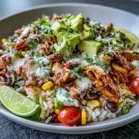 A colorful bowl of shredded salsa chicken over rice, topped with black beans, corn, and fresh avocado slices.  